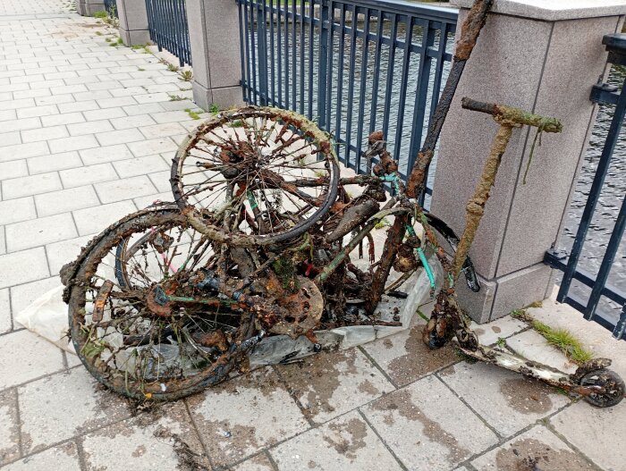 Rostiga cyklar och en elsparkcykel med skador, uppdragna från en å, ligger staplade intill ett räcke på en trottoar.