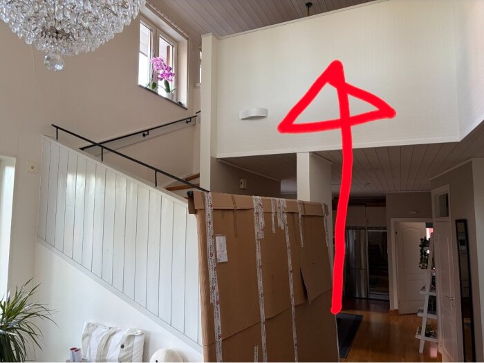 Stort paket vid trappa, röd pil pekar mot övre våning vid trappräcket, ljuskrona och fönster med blommor synliga.
