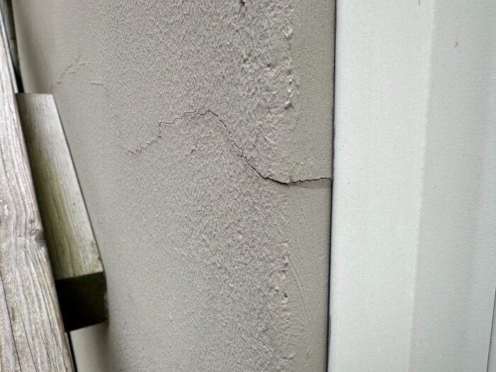 Spricka i utsidan av en putsad yttervägg, delvis igenspacklad, med en träbalk synlig i vänster kant av bilden.