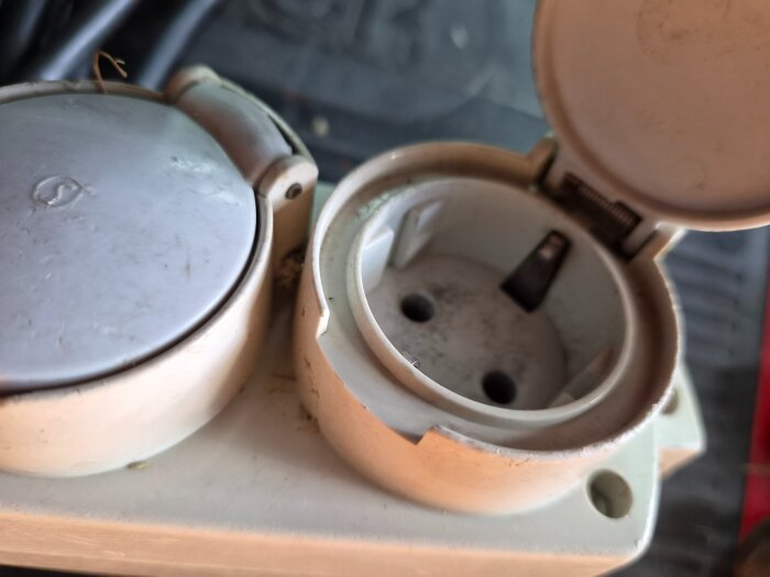 Vägguttag med upphöjd design och fläns, lock delvis öppet; skydd mot fukt, anpassat för utomhusbruk.