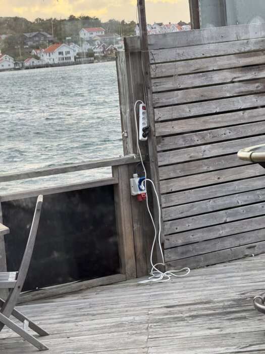 Eluttag och förlängningssladd installerade på en trävägg utomhus vid havet, med flera hus i bakgrunden och en del av ett bord synligt.