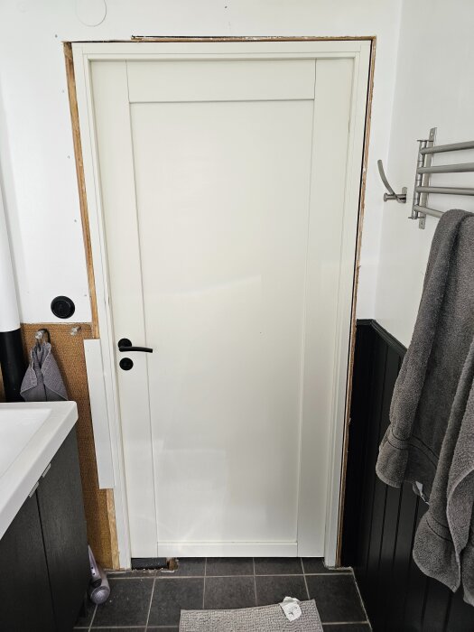 Vit dörr med karm monterad i en toalett, synlig nivåskillnad och mellanrum mellan karm och väggpanel som är målad och har svart bröstpanel nertill.