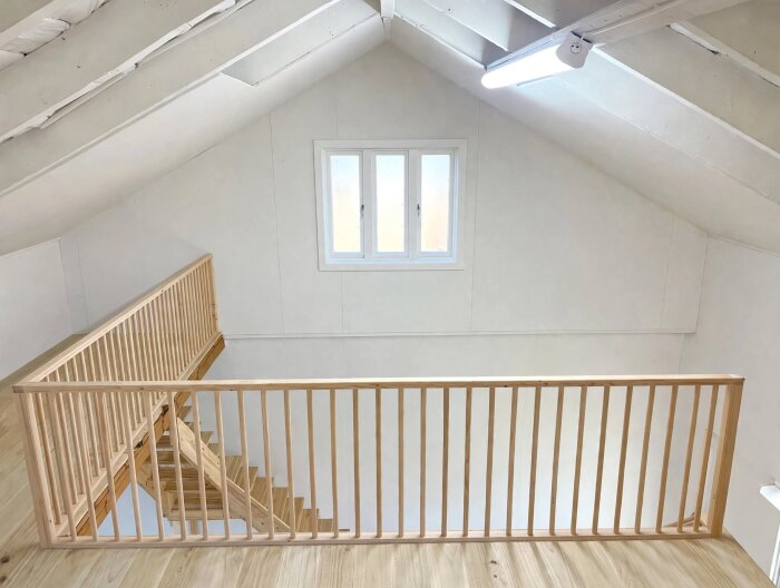 Ljusa loft med snedtak och ljuskälla, träräcke längs trappan, vitmålat tak och golv.