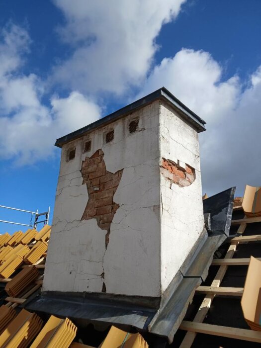 Skorsten på äldre hus med skadad puts och synligt tegel, omgiven av takpannor under reparation.