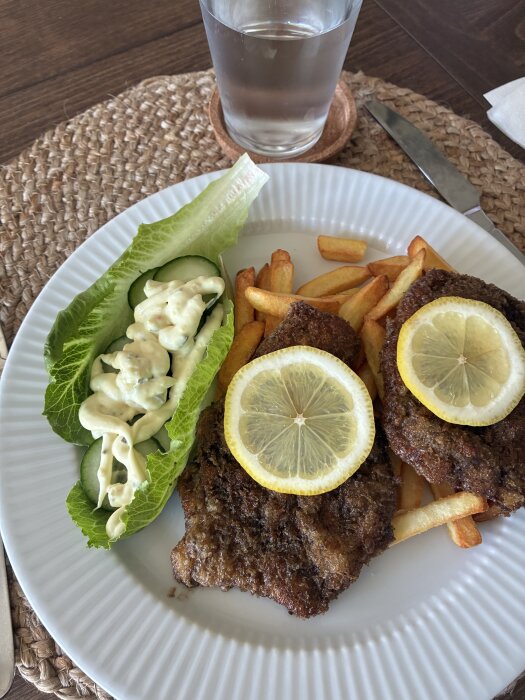 Panerad kotlett med citronskivor, pommes frites och sallad med gurka och remouladsås på en tallrik, bredvid ett glas vatten.