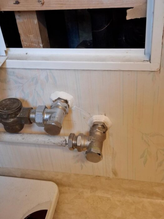 Rörkopplingar i ett badrum från 1982 med märkningen AHA, visar två rör med ventiler och vita lock, det övre rör för varmvatten och det nedre för kallvatten.