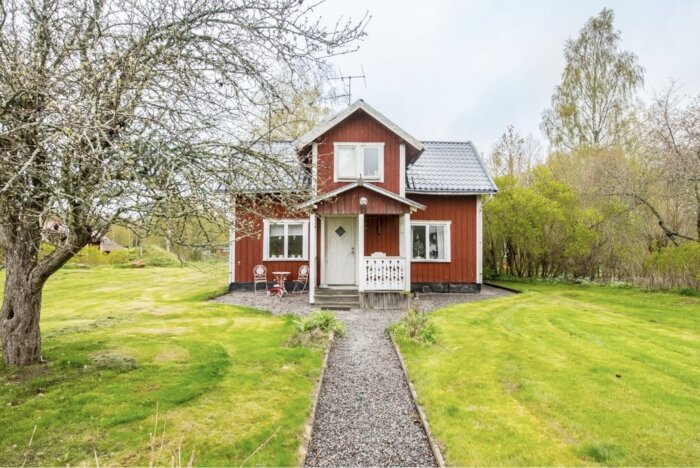 Rött trähus med vita knutar i en grönskande trädgård, synlig grusgång och äldre träd framför huset.