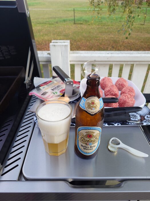 Flaska och glas med veteöl vid grill, omgiven av råa hamburgare och grilltillbehör.