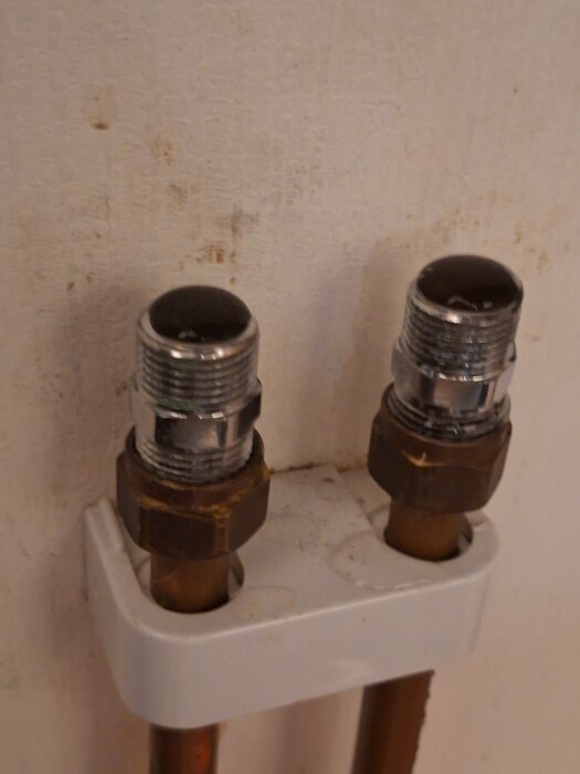 Två kopparrör med metallanslutningar är fästa vid en vägg, monterade genom en vit hållare.