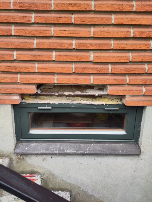 Tegelmur med delvis borttagna stenar ovanför källarfönster på hus från 1971, visar rostig armering och nyligen ommurat parti.