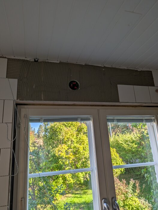 Hål i väggen ovanför kakel i kök, synligt genom stora fönster mot trädgård; inga överskåp planeras, endast hyllor.