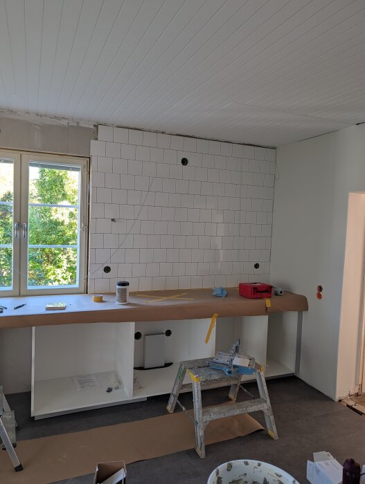 Vitt kakel på köksvägg med flera hål tagna i, verktyg och byggmaterial på bänkskiva, stege och fönster synliga.