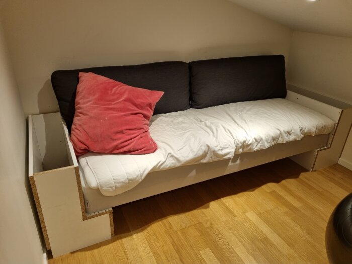 Hemmagjord soffa av en sängbotten med vita hyllplan som armstöd, svart och röd kudde, och liggunderlag på ett trägolv i ett hörnrum.