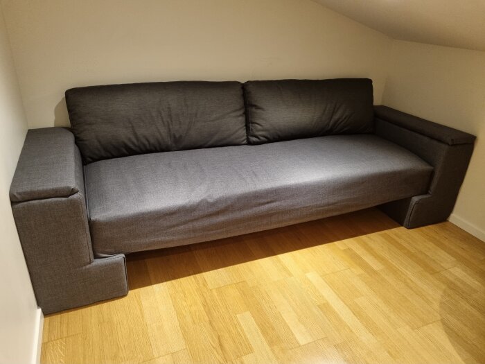 Grå soffa i ett vindsrum, med förvaringsbara armstöd för täcken och kuddar. Gjord från en gammal sängbotten och återvunna soffkuddar.