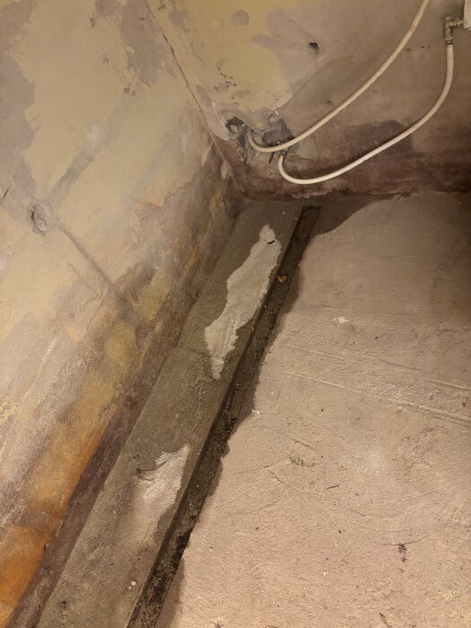 Betongkällare med fuktiga fogar och hörn visar tecken på vattensamling, med ledningar längs väggen. Potentiellt dräneringsproblem.
