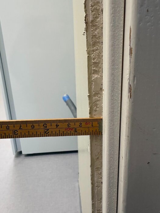 Närbild på en dörrkarm där gipsskivans kant avslutas ojämnt, med ett måttband som visar avståndet mellan dörrkarmen och väggen.