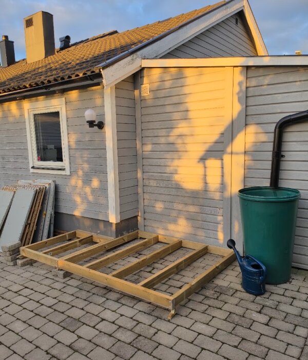 Träram på stenlagd yta vid husvägg, förberedd för förråd. Vattenkanna och tunna nära. Solnedgångens ljus skapar skuggor på fasaden.