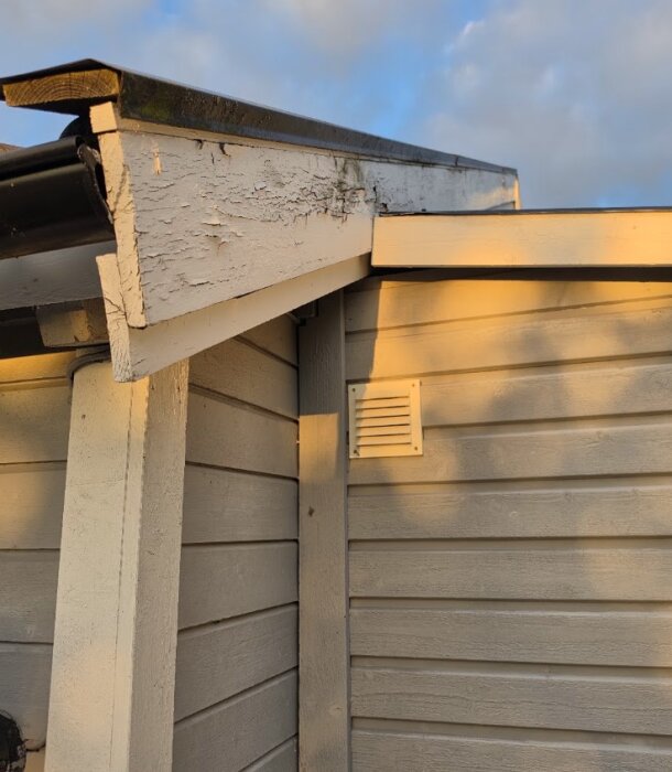 Knepigt hörn där takutsprång från större hus möter nybyggt förråd, med synligt lutande tak och bottenram, målade träpaneler.