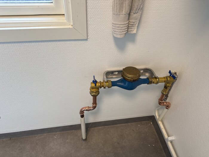 Vattenmätare och kopparrör monterade på en vitmålad vägg i ett rum med grått golv och ett fönster med fönsterbräda ovanför.