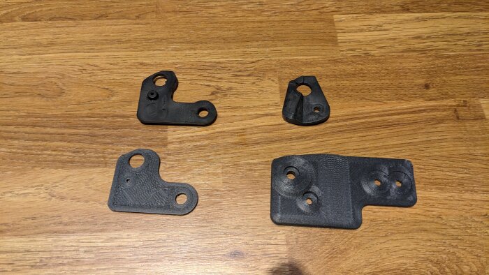 Svarta, 3D-printade förstärkningsbitar i plast för kylskåpsdörr och gångjärn, liggandes på träbord. Fyra olika former syns.