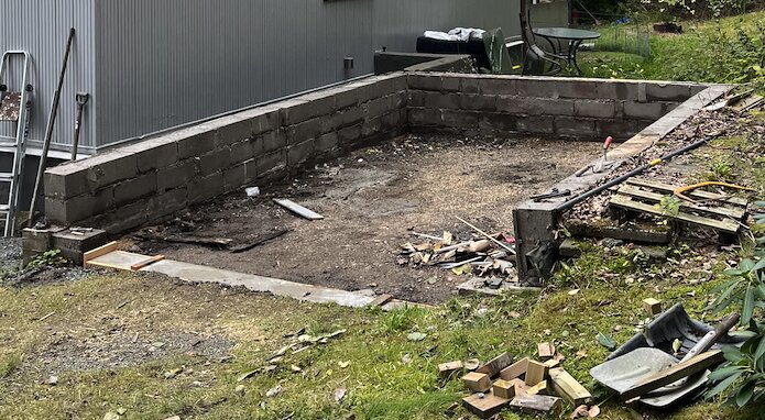 Grund av ett rivet garage med synliga betongblock och stenar. Ett område förberett för att bygga ett förråd. Verktyg och byggmaterial finns runtom.