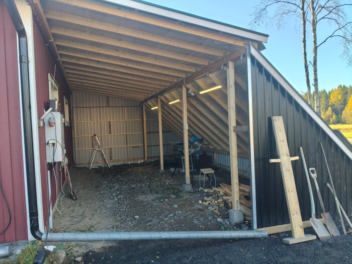 Halvbyggd carport med snedtak och LED-lysrör inuti. Spirorör vid sidan om för vattenavledning, med byggmaterial och verktyg på marken.