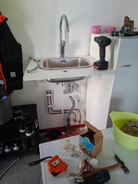 Diskbänk med verktyg, borrmaskin och vattenslang. Under diskbänken finns rör och en dräneringsslang. I förgrunden skruvar och verktyg.