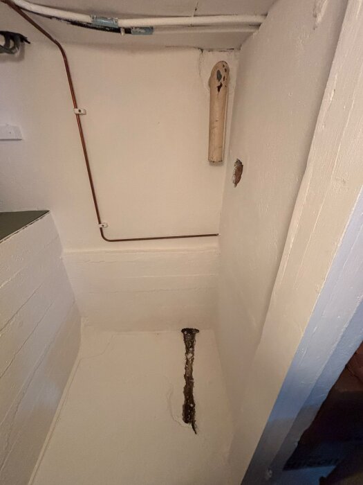 Litet rum i källare med dike i golvet och rör synligt på väggen. Diket sträcker sig från väggen och under golvplattan.