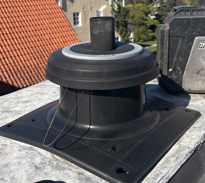 Svart ventilationshuv på skorstenstopp, med omgivande tak och grönska i bakgrunden. Frågeställning om funktion och behov av åtgärd.