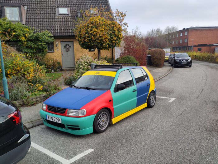 Flerfärgad bil parkerad på gata framför ett hus med trädgård och tegelhus i bakgrunden.