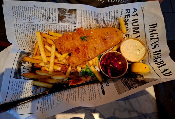 Friterad fisk med pommes frites på tidningspapper, serverad med sås, rödkål och en citronskiva.