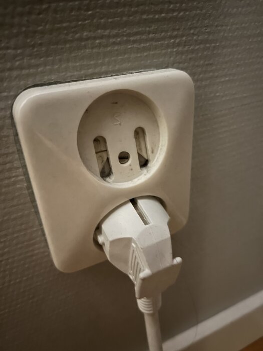 Ojordad väggkontakt i äldre stil med en ansluten vit sladd, monterad på en vägg med strukturerad yta.