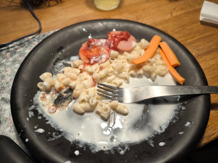 Svart tallrik med stuvade makaroner, falukorv och morotsstavar, tillsammans med en gaffel och en stor mängd sås.