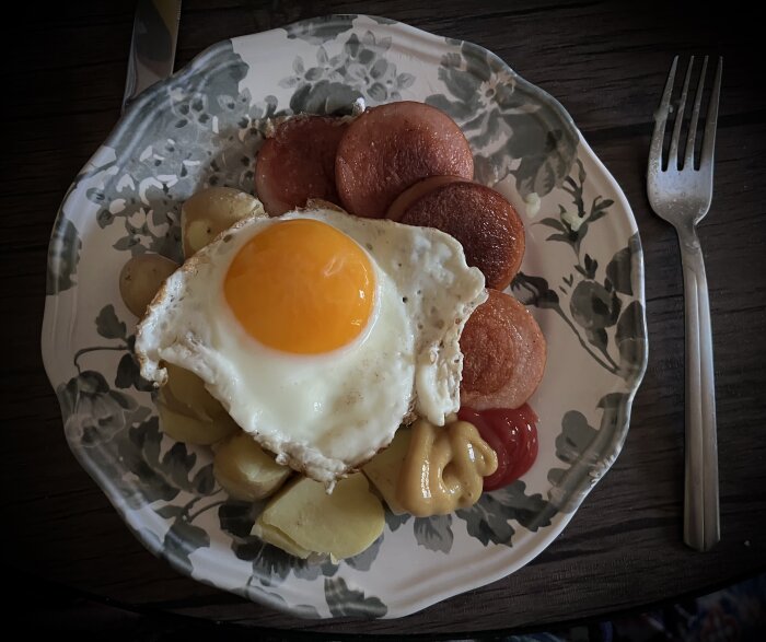 Stekt falukorv och ägg med kokt potatis, ketchup och senap på blommig tallrik.