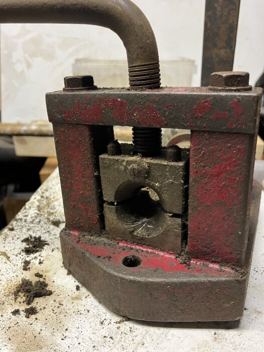 En gammal, sliten röd metallsvarv med rostiga skruvar och smutsig yta, placerad på en arbetsbänk med träskivor i bakgrunden.