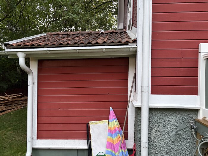 Röd husfasad med vita detaljer och sned lutning på träpanel samt stuprör. Tillbyggnadens tak lutar något och det finns en färgglad duk nedanför.