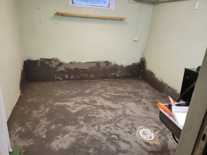 Tvättstuga med nyligen flytspacklat golv och uppmålad vägg som visar pågående renovering, med ett avlopp synligt i golvet.