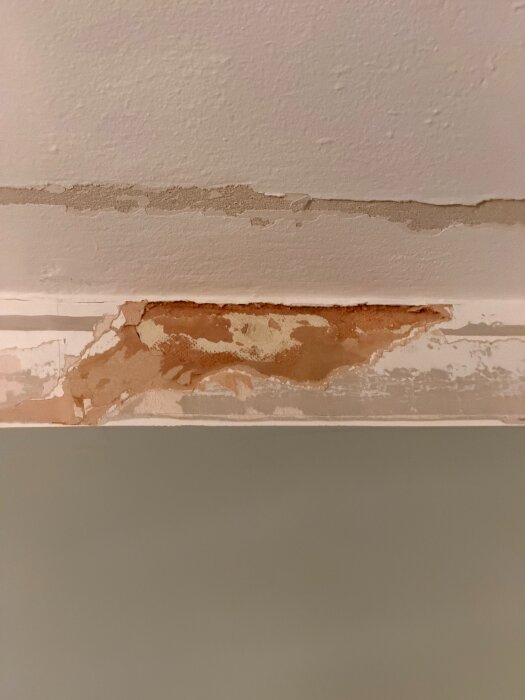 Skadad vägg och tak efter borttagning av frigolitlist med färg och underlag som följt med, vilket exponerar masonit och orsakar nivåskillnader.