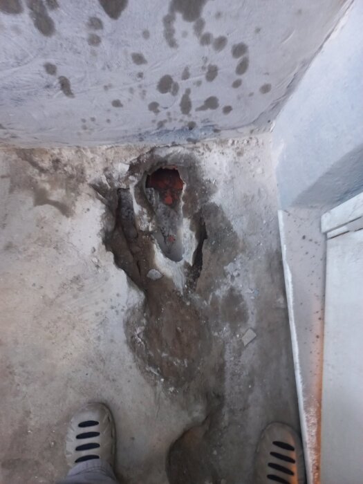 Gjutjärnsspillbrunn i betonggolv, omgiven av trasig betong och smuts vid en vägg. Slitage tydligt, närbild från källarrenovering.