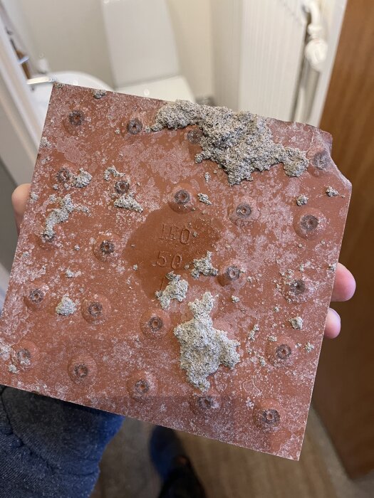 Baksida av en röd klinkerplatta med måtten 14,9x14,9 cm, lite trasig och täckt av cement eller murbruk, från renovering av ett radhus från 1951.