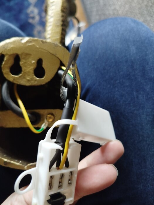 Närbild av vägglampa i metall med synliga kablar och kopplingsbox, där en jordsladd fästs vid metallhöljet. Lampan ligger på ett knä.