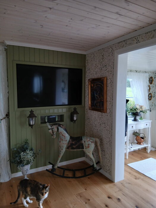 Enkelrum med vintage gunghäst, väggmonterad TV, inramad tavla och en katt på mörkt trägolv. Blommor i vas och vit byrå syns i bakgrunden.