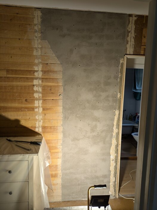 Murstock och borttagna gipsskivor syns på vägg. Verktyg och möbler täckta med plast vid renovering av ett äldre hus.