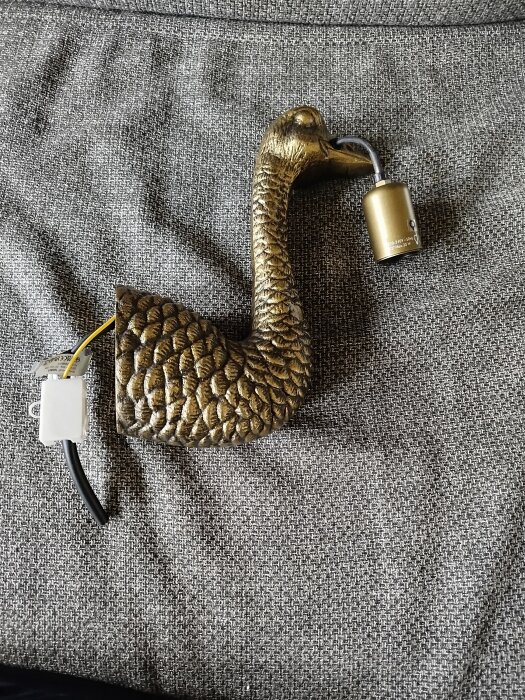 Guldmålad dekorativ svanfigur med ljushållare i näbben, liggandes på grått tyg.