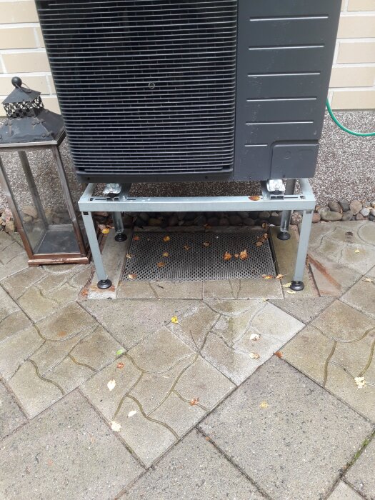 Utomhus installerad värmepump från Panasonic K-serie på metallställning, omgiven av stenar och betongplattor med en lykta bredvid.