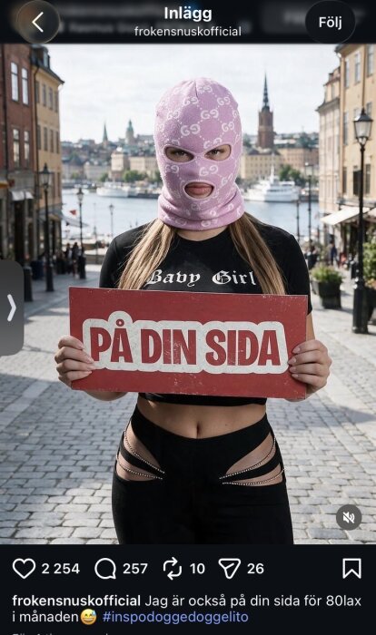 En person i rosa balaklava med Gucci-logotyp håller en skylt med texten "PÅ DIN SIDA" framför en stadsmiljö med vatten i bakgrunden.