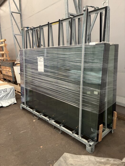 Inplastade glasrutor placerade på ett metallställ i en lagerlokal, redo för transport eller installation.