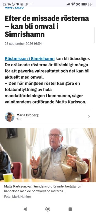 En man sitter i en fotölj, gestikulerar och talar. Bakgrunden visar ett ljust rum med gardiner.