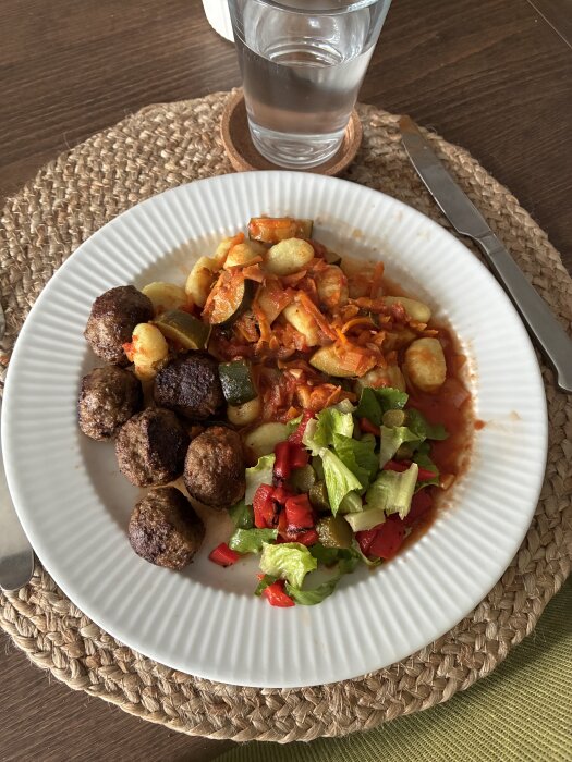 Tallrik med gnocchi i tomatsås arrabiata, köttbullar och sallad med grillad paprika och gröna blad, ett vattenglas i bakgrunden.
