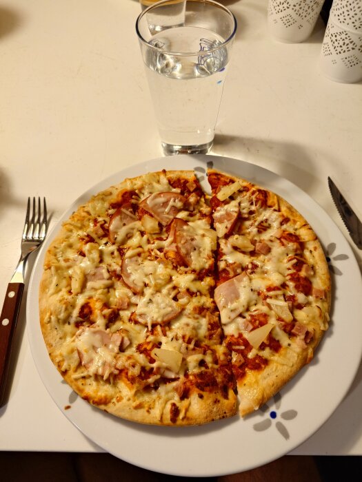 Stenugnsbakad Hawaii-pizza med skinka och ananas serverad på vit tallrik med glas vatten, gaffel och kniv bredvid.